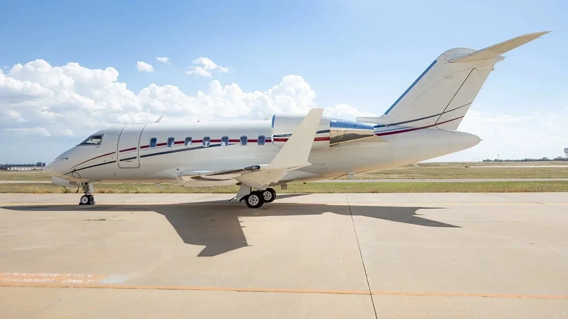 A white Bombardier Challenger business jet with red and blue stripes on the tail and fuselage, parked on an airfield under a partly cloudy sky.