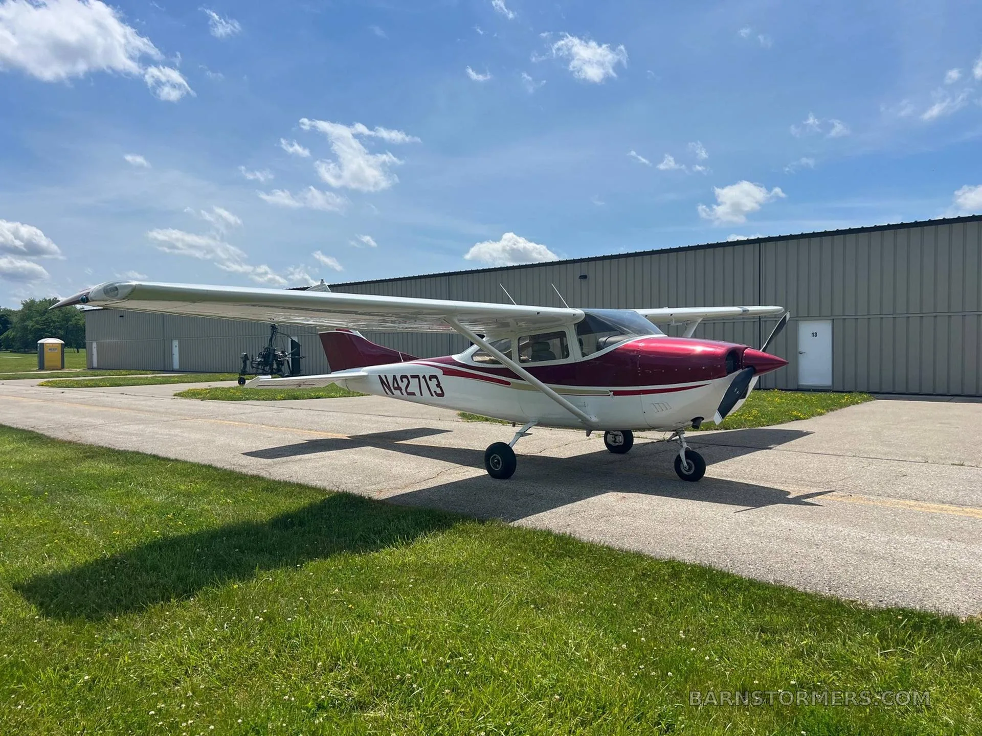 A red and white Cessna N42713 parked on an asphalt taxiway next to a hangar under a partly cloudy sky.