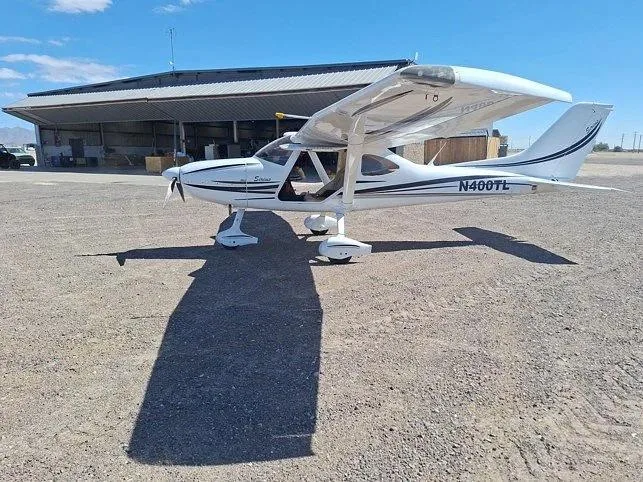 A white Sirius LightSport aircraft with black and grey stripes, registration N400TL, parked on a gravel apron in front of a hangar.