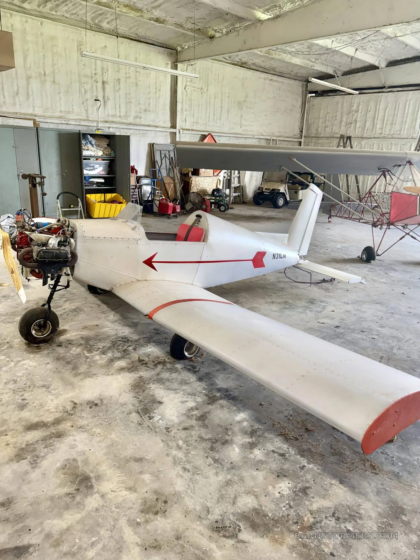 A white single-engine experimental aircraft with a red stripe and exposed engine, parked in a hangar.