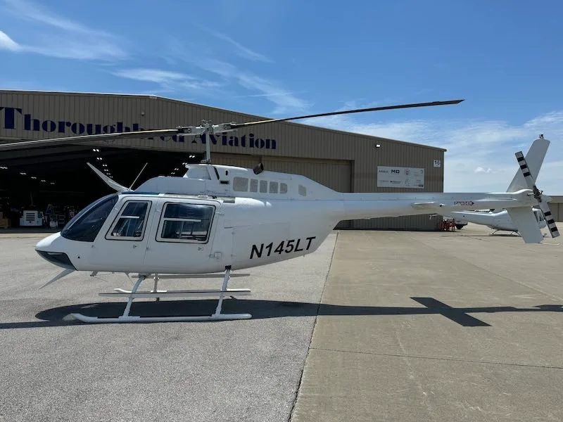 Side view of a white Bell 206L-4 helicopter with tail number N145LT in front of a hangar.
