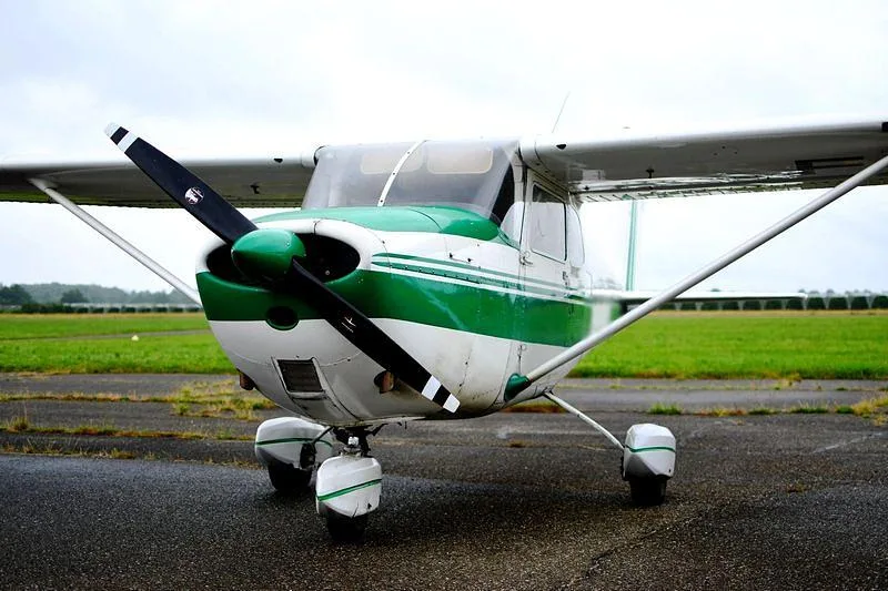 A white and green single-engine propeller aircraft, resembling a Cessna, parked on a tarmac.