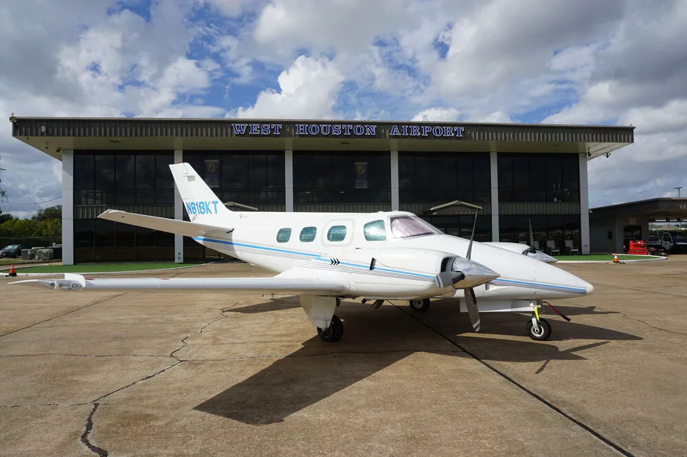 A white twin-engine propeller aircraft with blue stripes, registration N818KT, parked on the tarmac with the West Houston Airport building behind it.