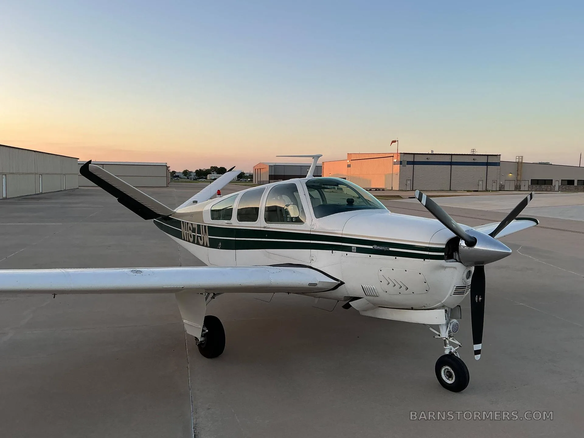 A front-quarter view of a white and dark green Beechcraft Bonanza (N167JW) parked on an airfield tarmac at sunset.