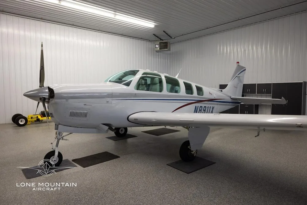 A white Beechcraft Bonanza A36 aircraft with blue and red stripes, parked in a hangar.