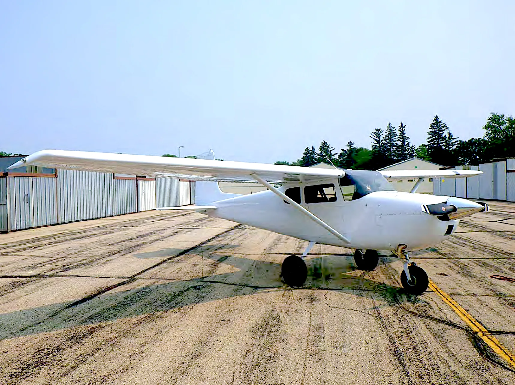A white single-engine high-wing aircraft is parked on an asphalt ramp next to hangars under a clear sky.
