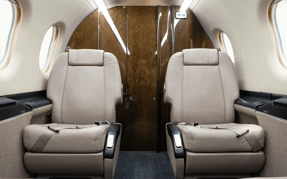 Two beige leather passenger seats in the cabin of a Pilatus PC-12 NG, facing a wood-paneled bulkhead.