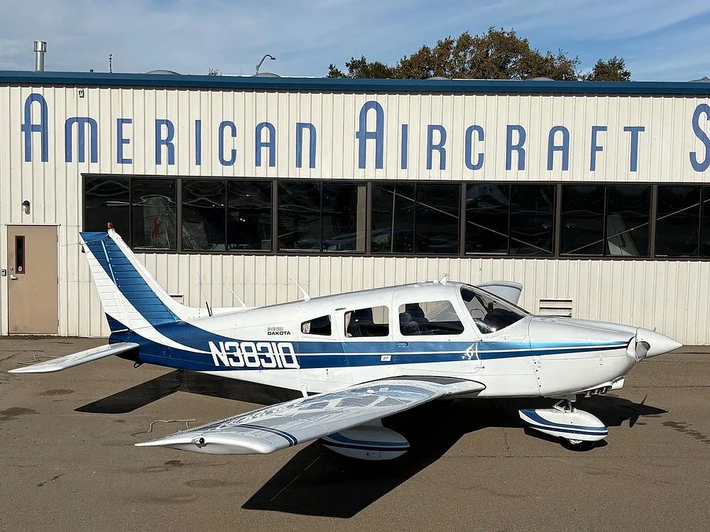 A white Piper Dakota aircraft with blue stripes, registration N3831Q, parked in front of a building labeled "American Aircraft Sales".