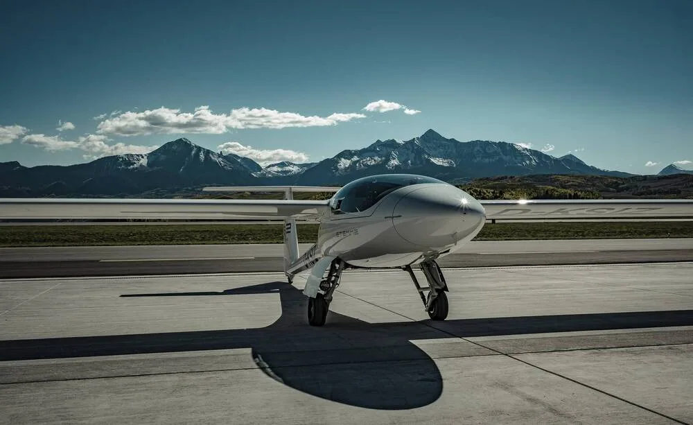 A white Stemme S12 motorglider with registration D-KSIZ parked on an airfield with snow-capped mountains in the background.