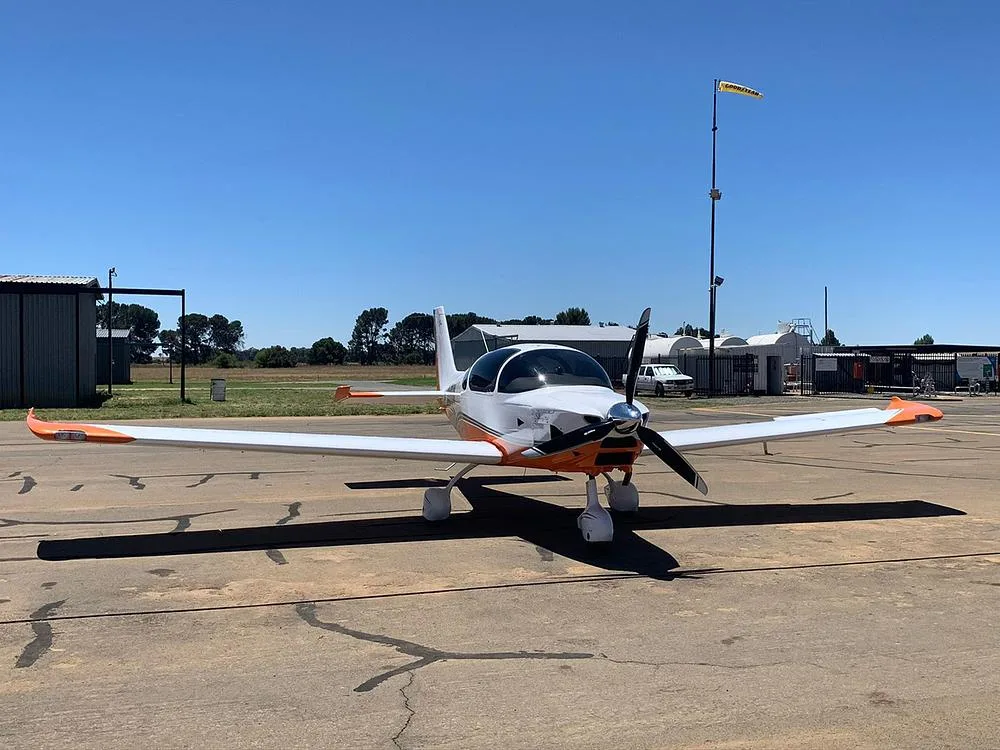 A white and orange light aircraft with a propeller sits on an asphalt apron under a clear blue sky.