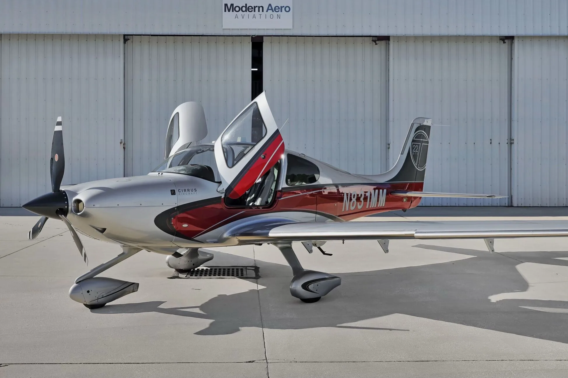 Side view of a Cirrus SR22T aircraft with red and silver paint scheme, registration N831MM, parked on an airfield with doors open.