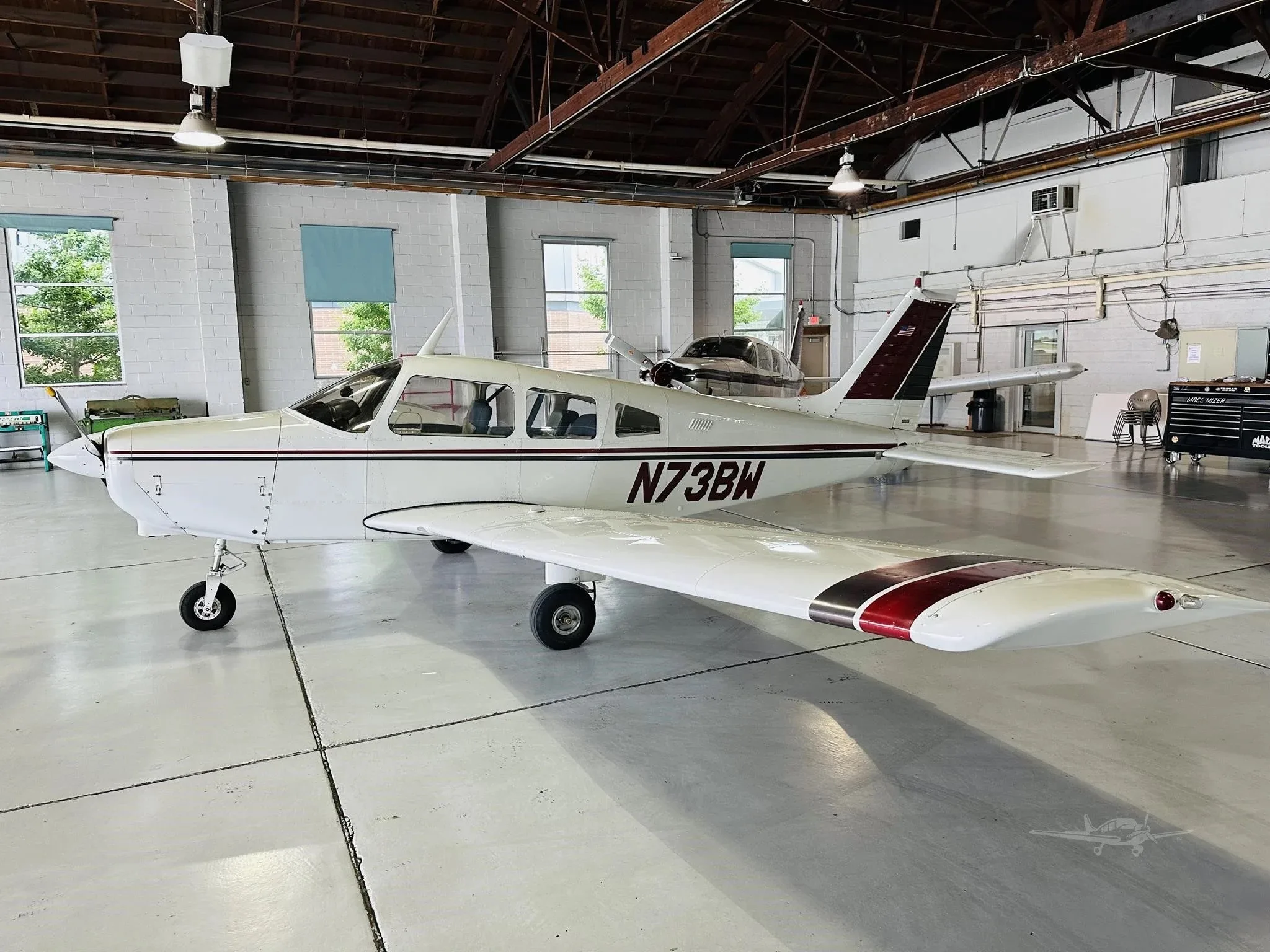 A white single-engine aircraft with maroon and black stripes, registration N73BW, viewed from the left side in a hangar.
