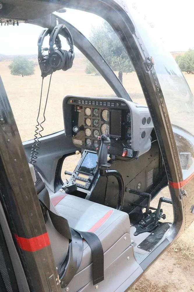 The pilot's side of a helicopter cockpit features the instrument panel, controls, and a grey and red seat with headphones hanging.