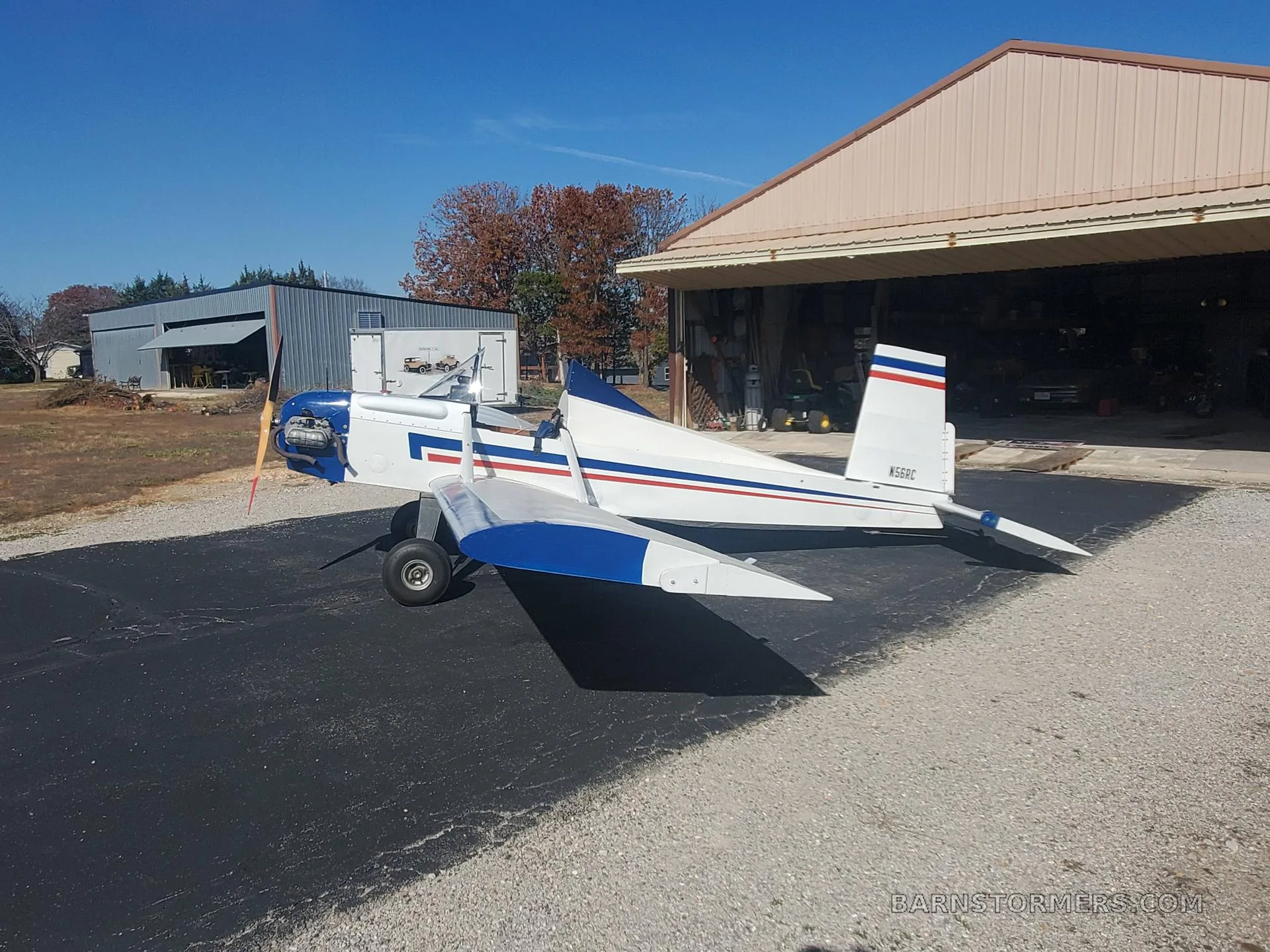 A white and blue single-engine aircraft with a red propeller tip is parked on asphalt next to an open hangar under a clear blue sky.
