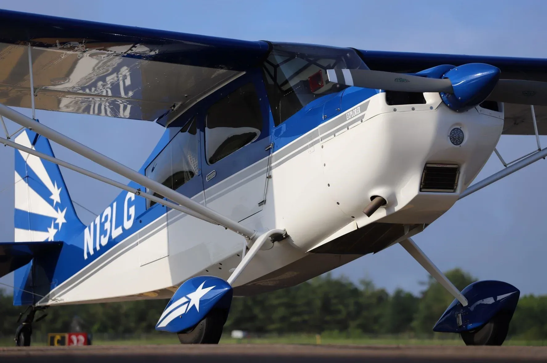 A blue and white high-wing monoplane, tail number N13LG, with star decals, seen from a low front-right angle on an airfield.