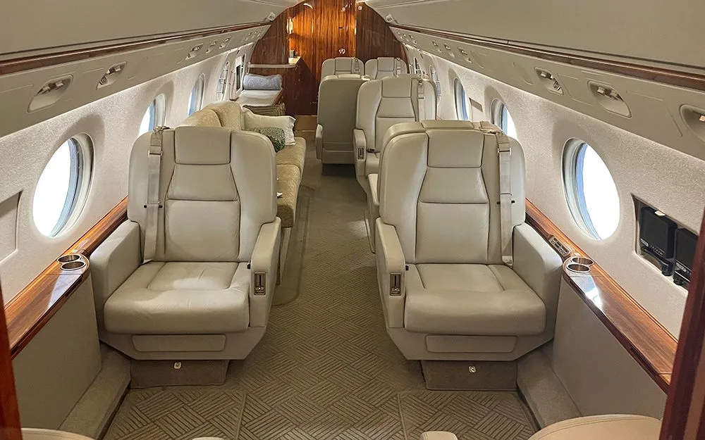 A wider view of the private jet's cabin, showing rows of light beige leather seats, large windows, and polished wood accents.