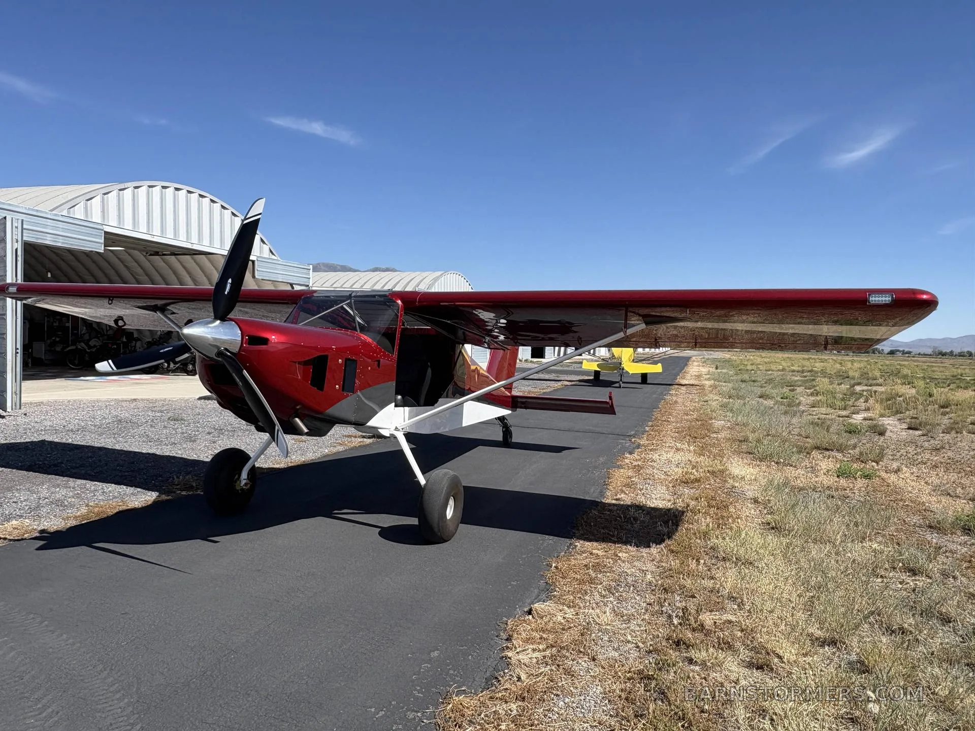 A red and white single-engine aircraft with a propeller on an asphalt strip in front of hangars under a clear blue sky.