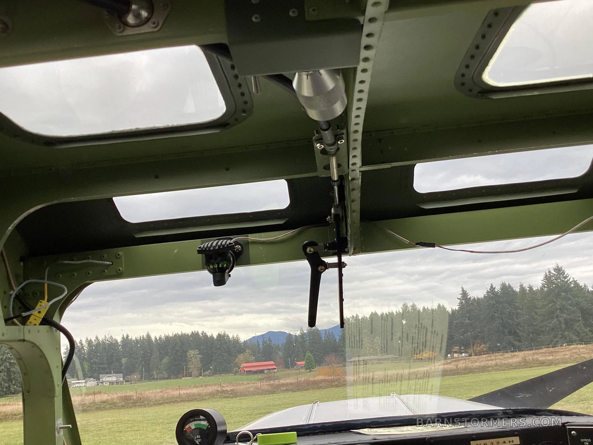 View from inside the cockpit of a floatplane, looking out over a grassy field and trees, showing the overhead panel and a control stick.