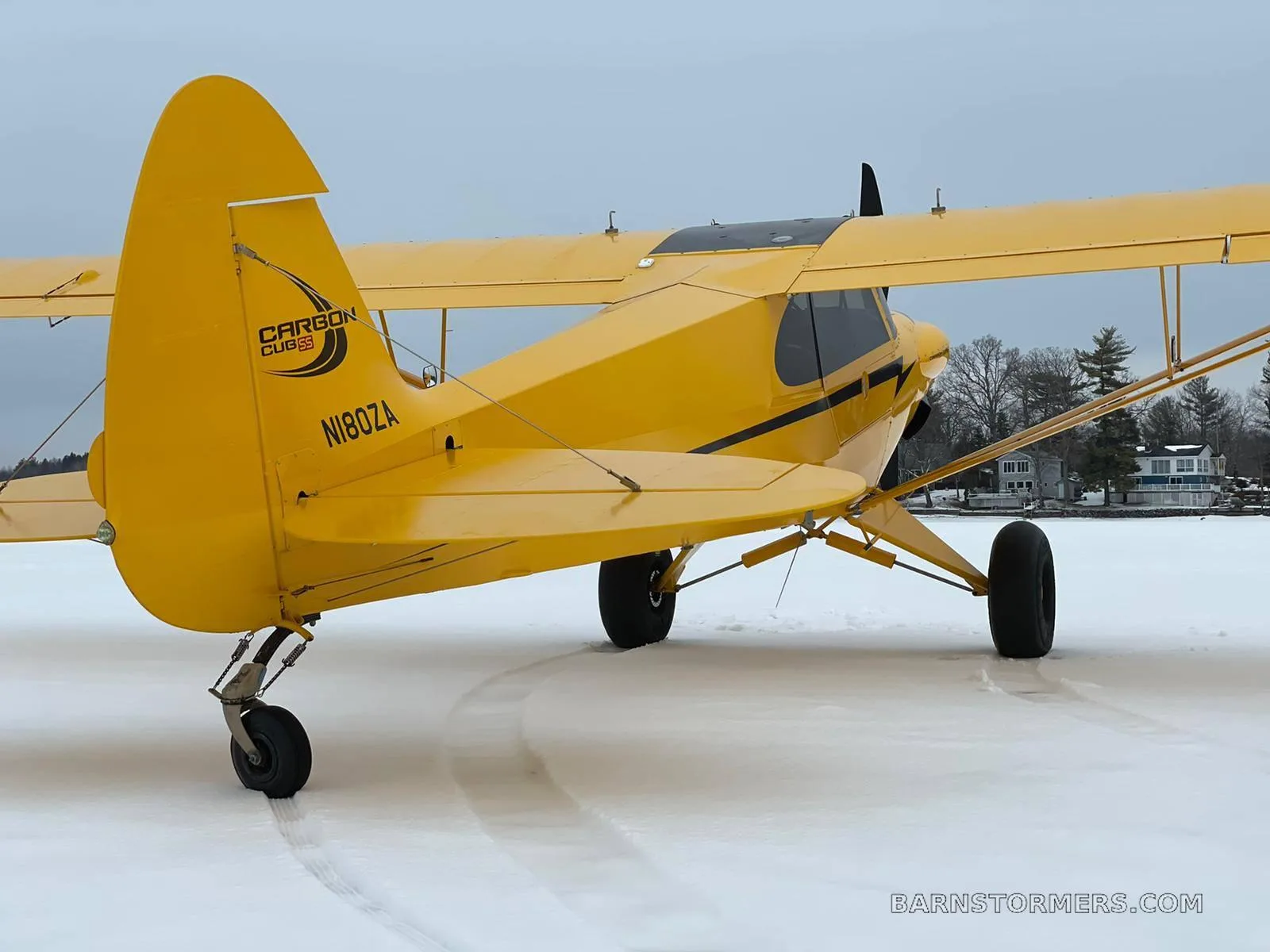 A yellow Carbon Cub aircraft with tail number N180ZA is parked on a snowy surface next to a lake.
