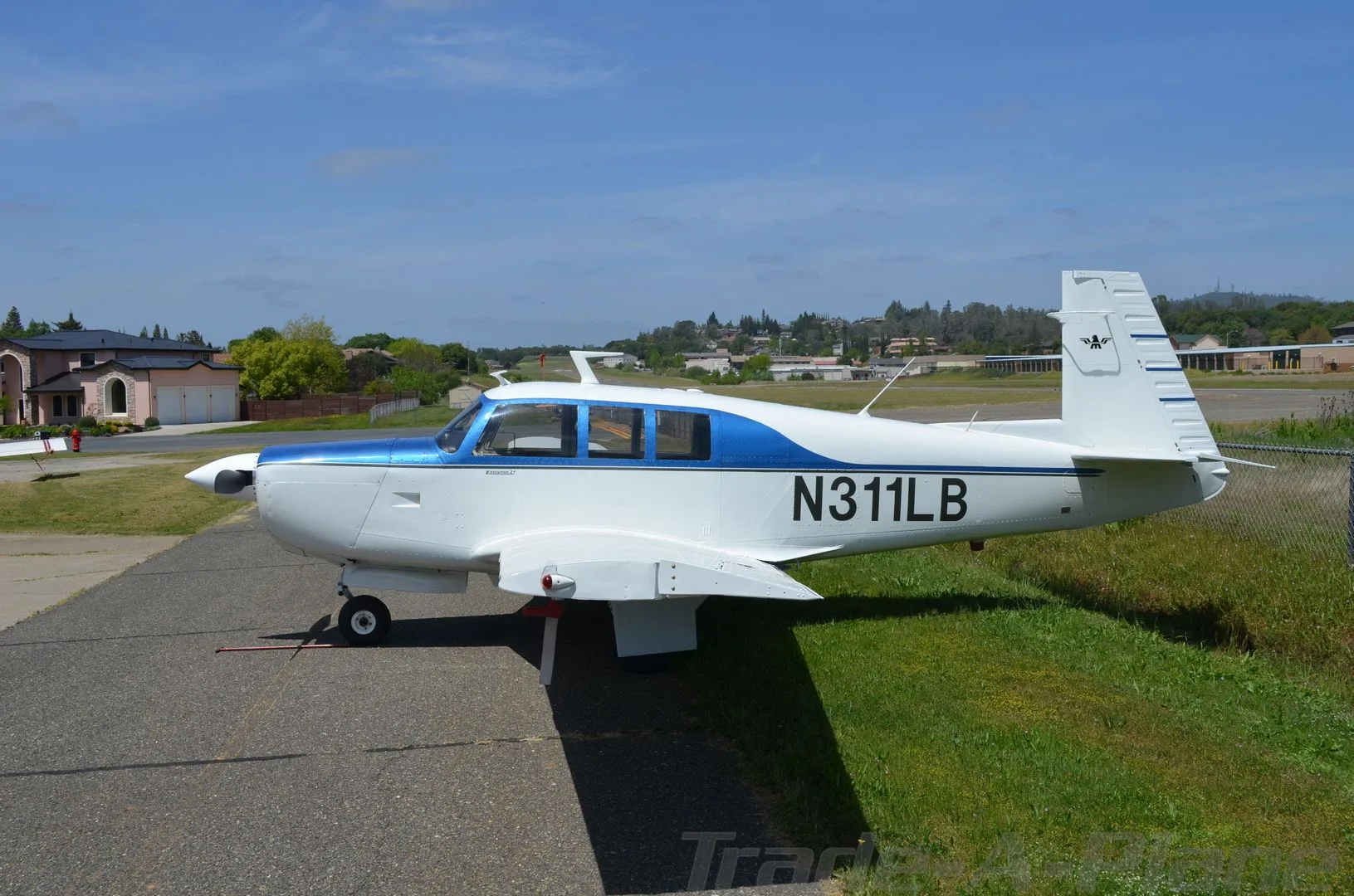 A white and blue single-engine propeller aircraft, registration N311LB, is parked on an asphalt ramp next to a grassy area under a clear sky.