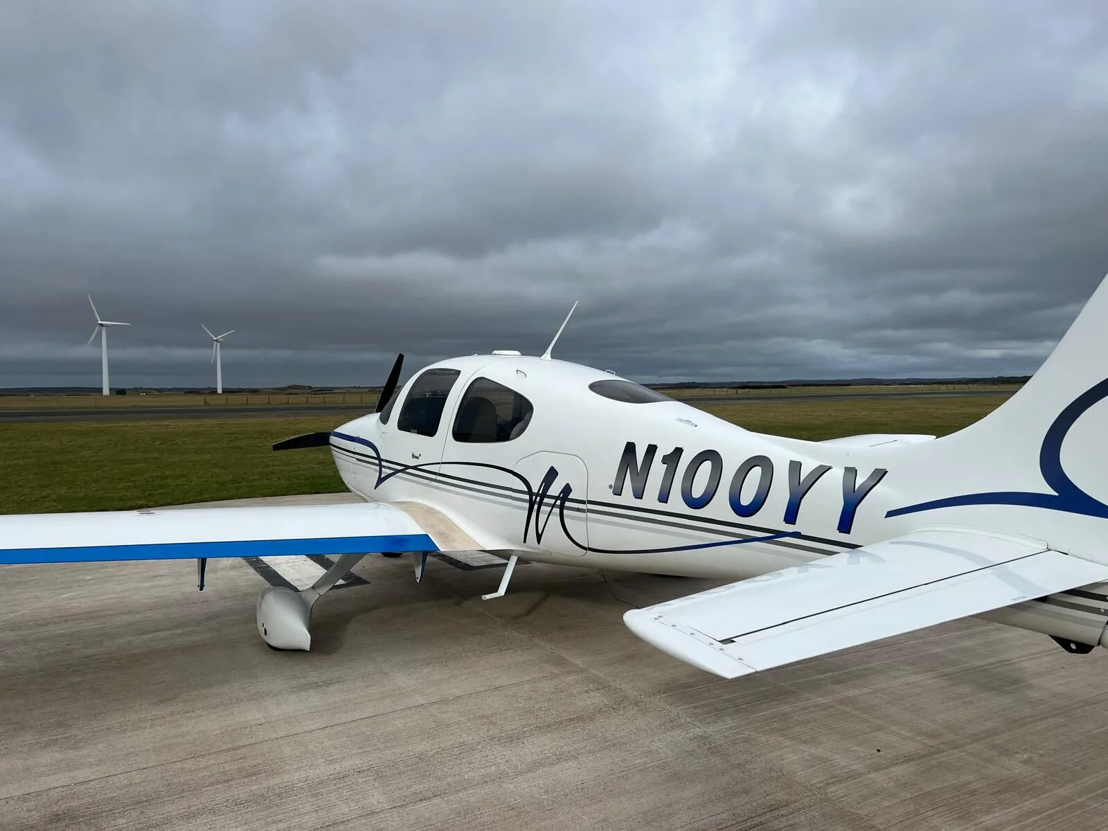 A white Cirrus SR22 aircraft with blue and grey stripes parked on an airfield under a cloudy sky, with wind turbines in the distance.