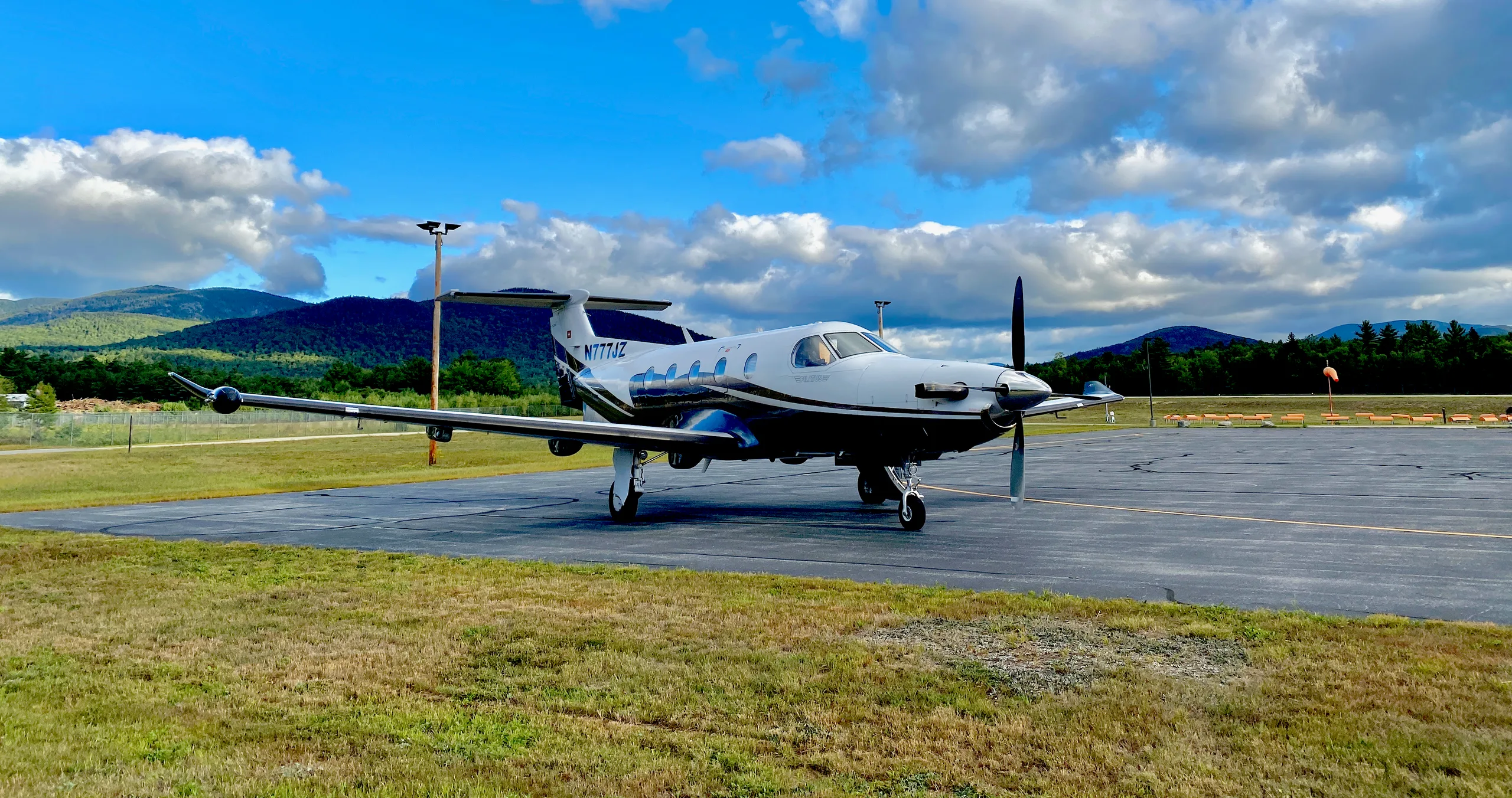 A Pilatus PC-12 aircraft with registration N777JZ is parked on the tarmac with mountains in the background.