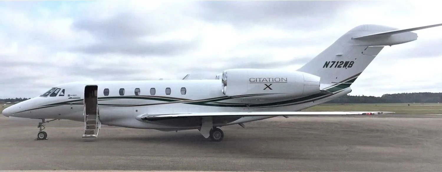 A white Cessna Citation X business jet with green and black stripes, registration N712WB, parked on an airfield with its main door open.