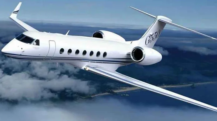 A white Gulfstream G550 business jet with blue stripes flies above clouds and a coastline.