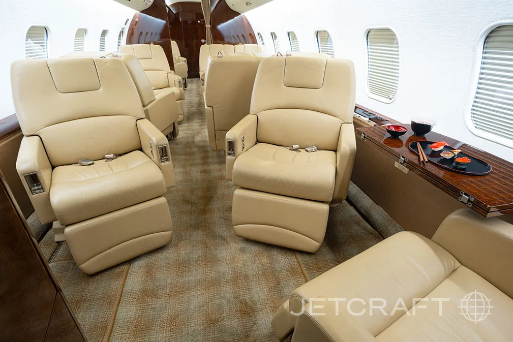 A private jet cabin interior featuring beige leather seats, a side table with sushi, and a patterned carpet.