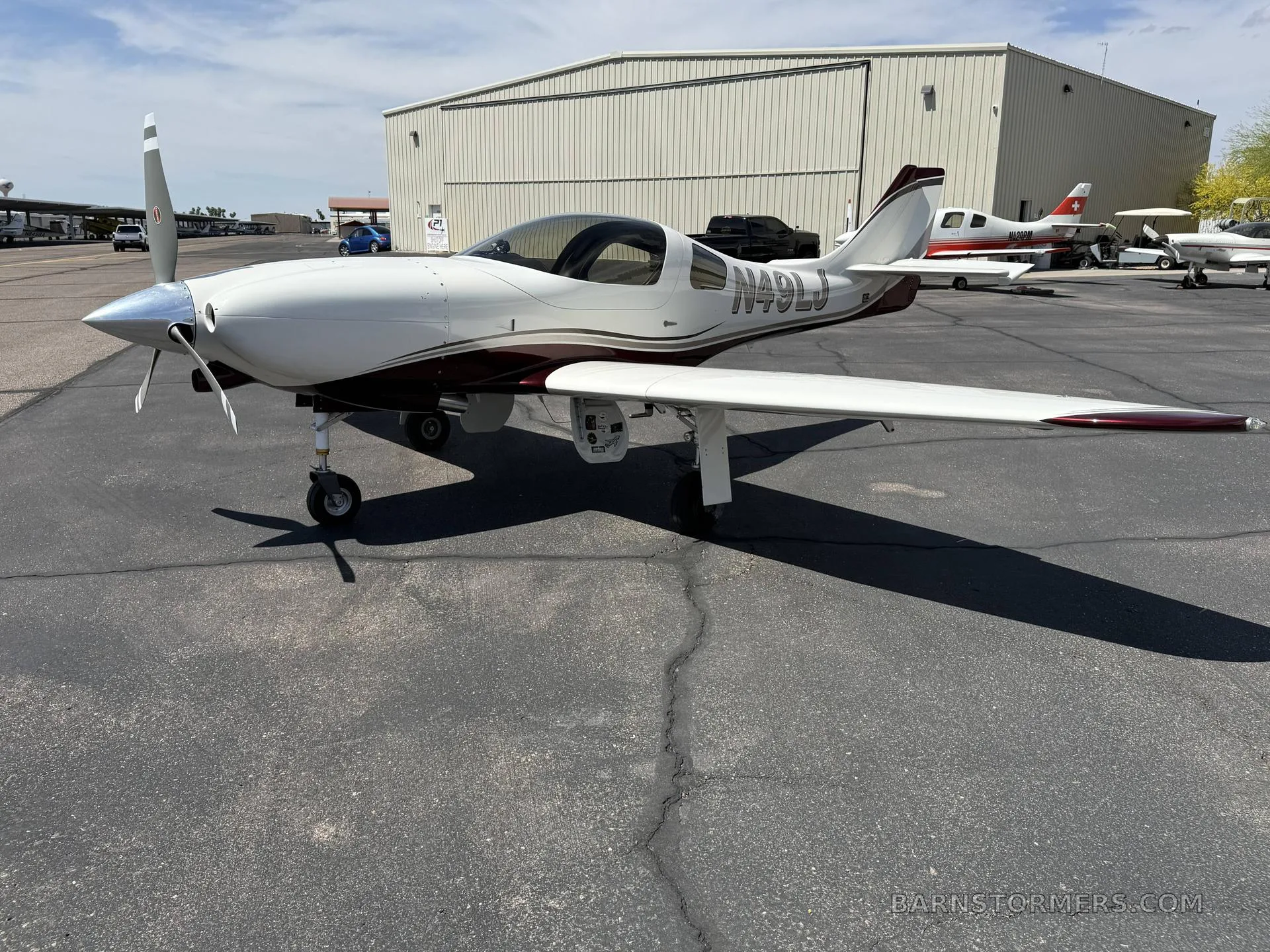 A white and maroon single-engine aircraft (N49LJ) with a propeller, parked on an asphalt apron with a hangar and other aircraft in the background.