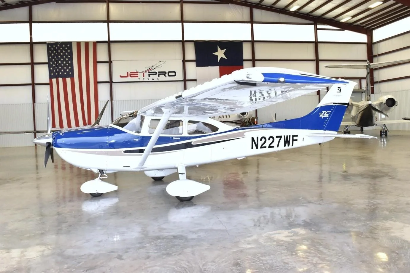 A white and blue Cessna Skylane N227WF propeller aircraft parked inside a hangar with flags.