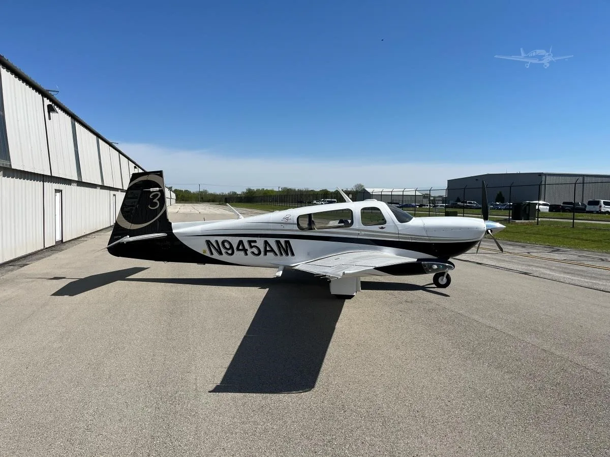A white and black single-engine propeller aircraft (N945AM) with gold accents is parked on an apron next to a hangar under a clear blue sky.