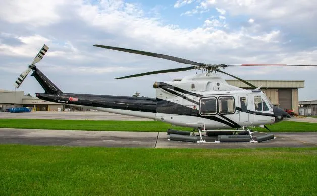 A white and black helicopter with floats is parked on an asphalt pad with grass, under a cloudy sky.