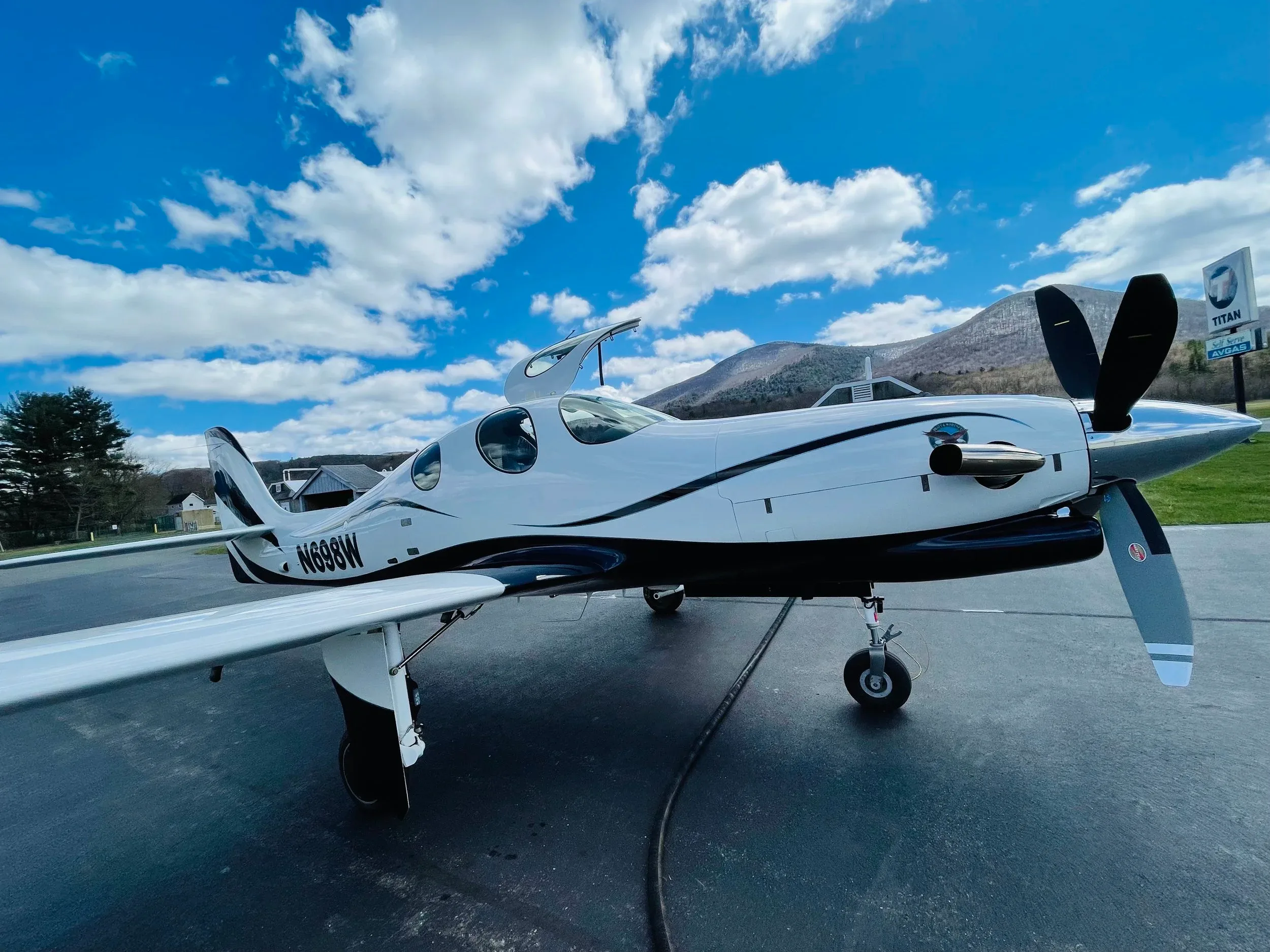 A white and black single-engine turboprop aircraft, registration N698W, is parked on an airfield with its doors open, against a backdrop of mountains and a blue sky with clouds.
