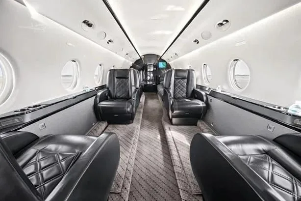 The luxurious interior of a private jet cabin featuring black leather seats in a club configuration and a patterned aisle carpet.