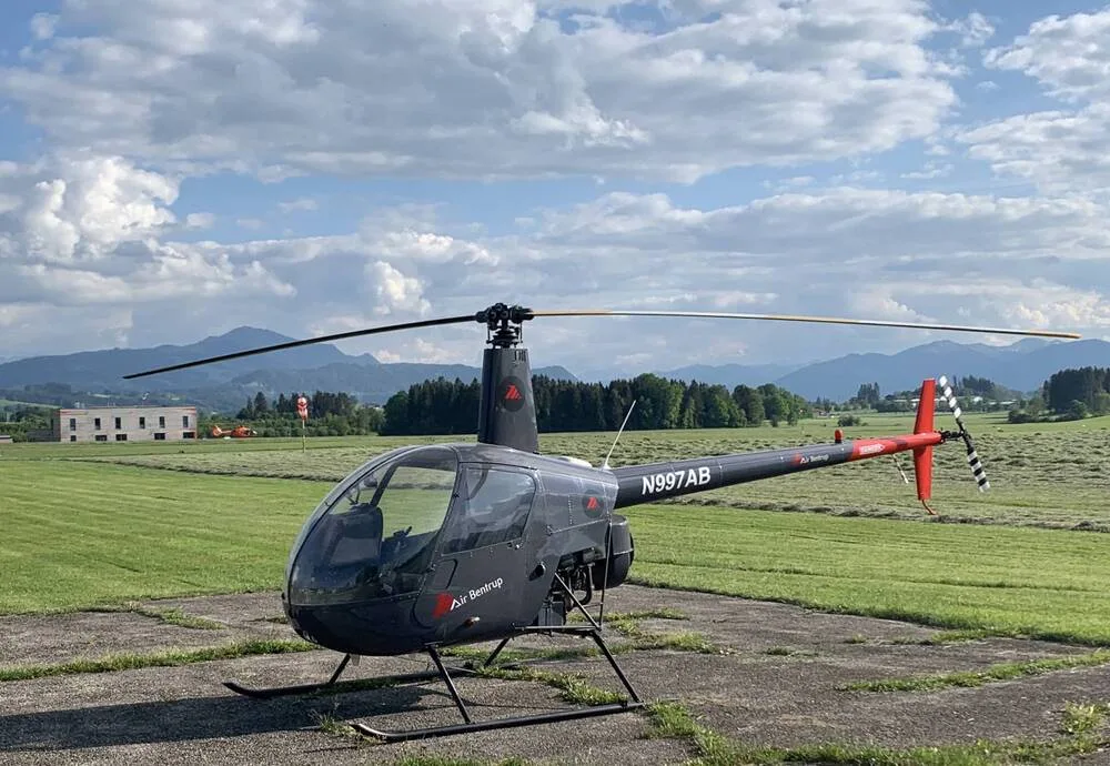 A dark gray Robinson R44 helicopter, N997AB, with red tail accents, parked on a paved area next to a field with mountains in the distance.