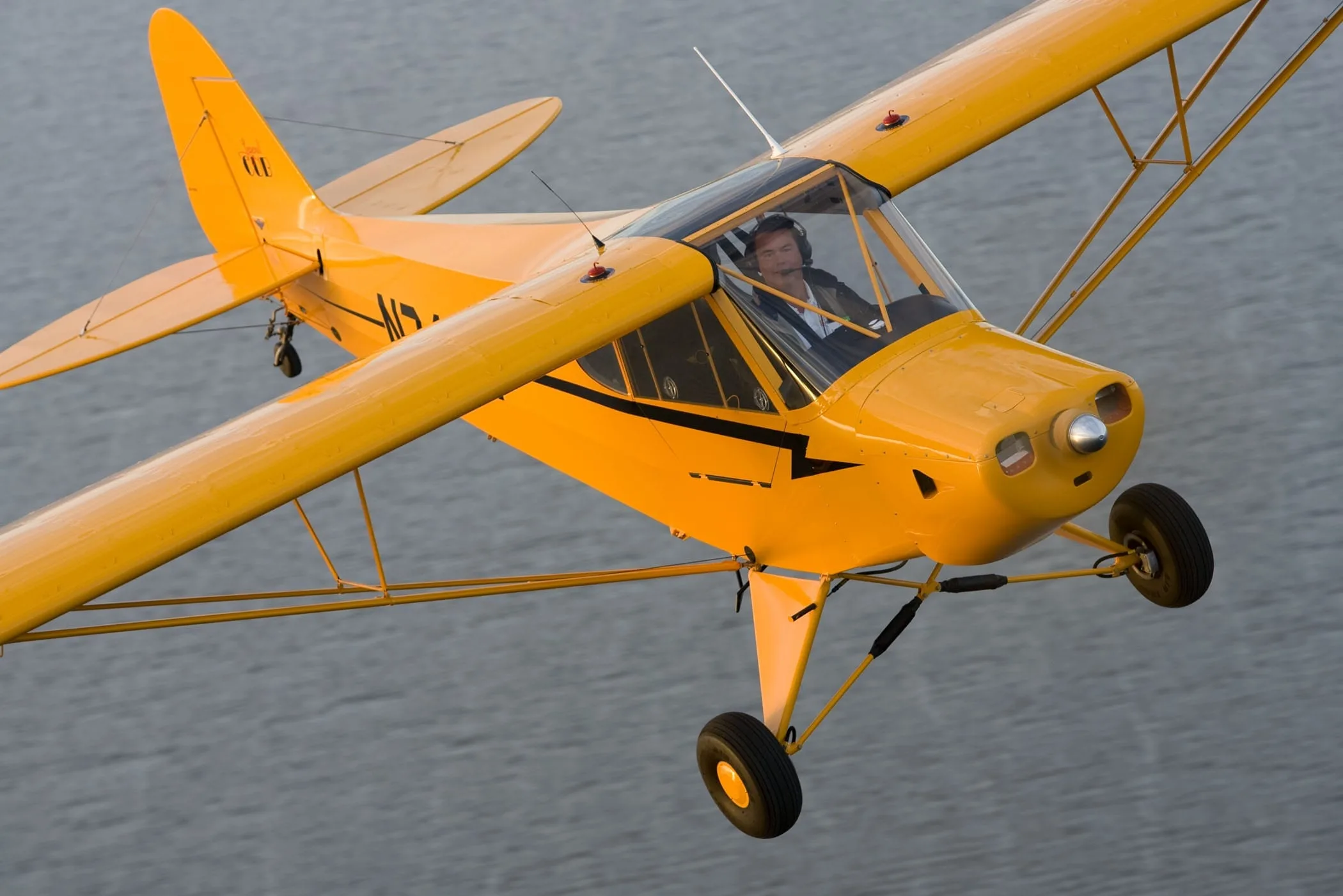 A yellow Piper Cub-style aircraft (N741W) with a pilot visible in the cockpit flies over water.