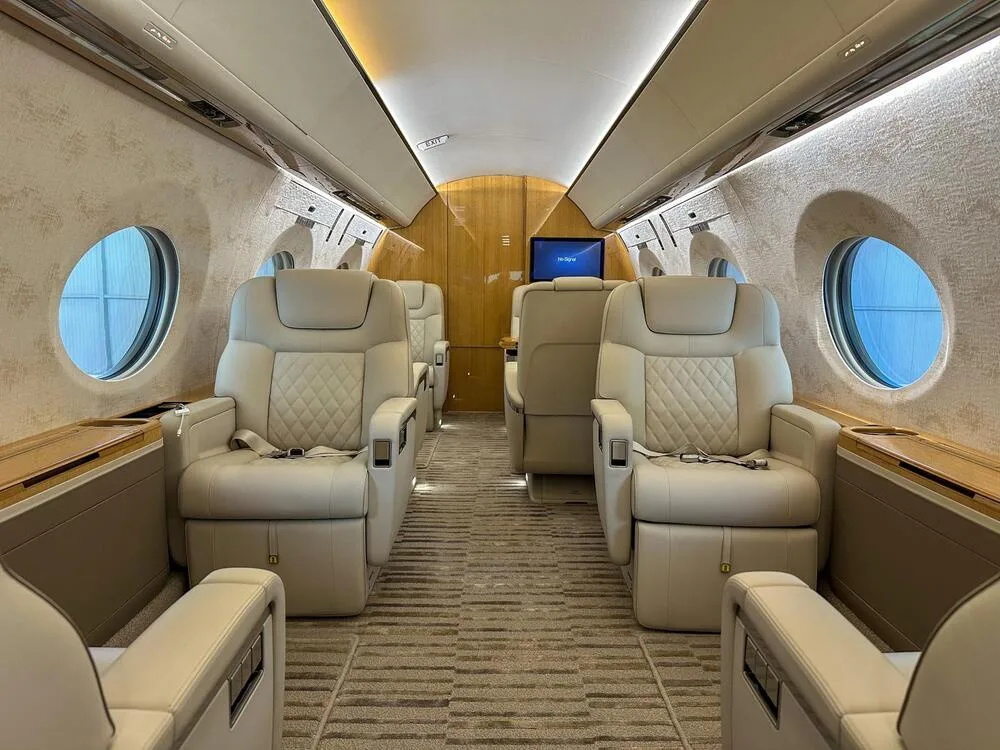 A wide shot of a luxurious aircraft cabin featuring light-colored quilted leather seats, wood accents, and large oval windows.