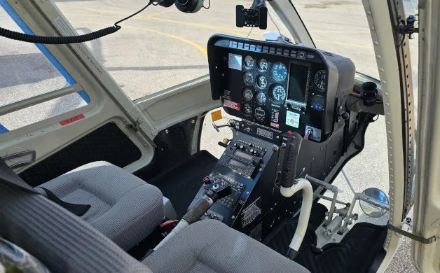 The cockpit of a helicopter shows the instrument panel, controls, and two grey fabric seats.