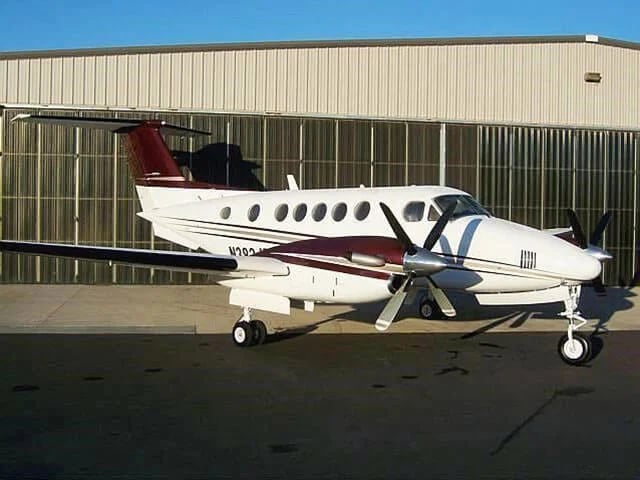 A white twin-engine turboprop aircraft with a maroon tail and stripes is parked on an airfield in front of a hangar.