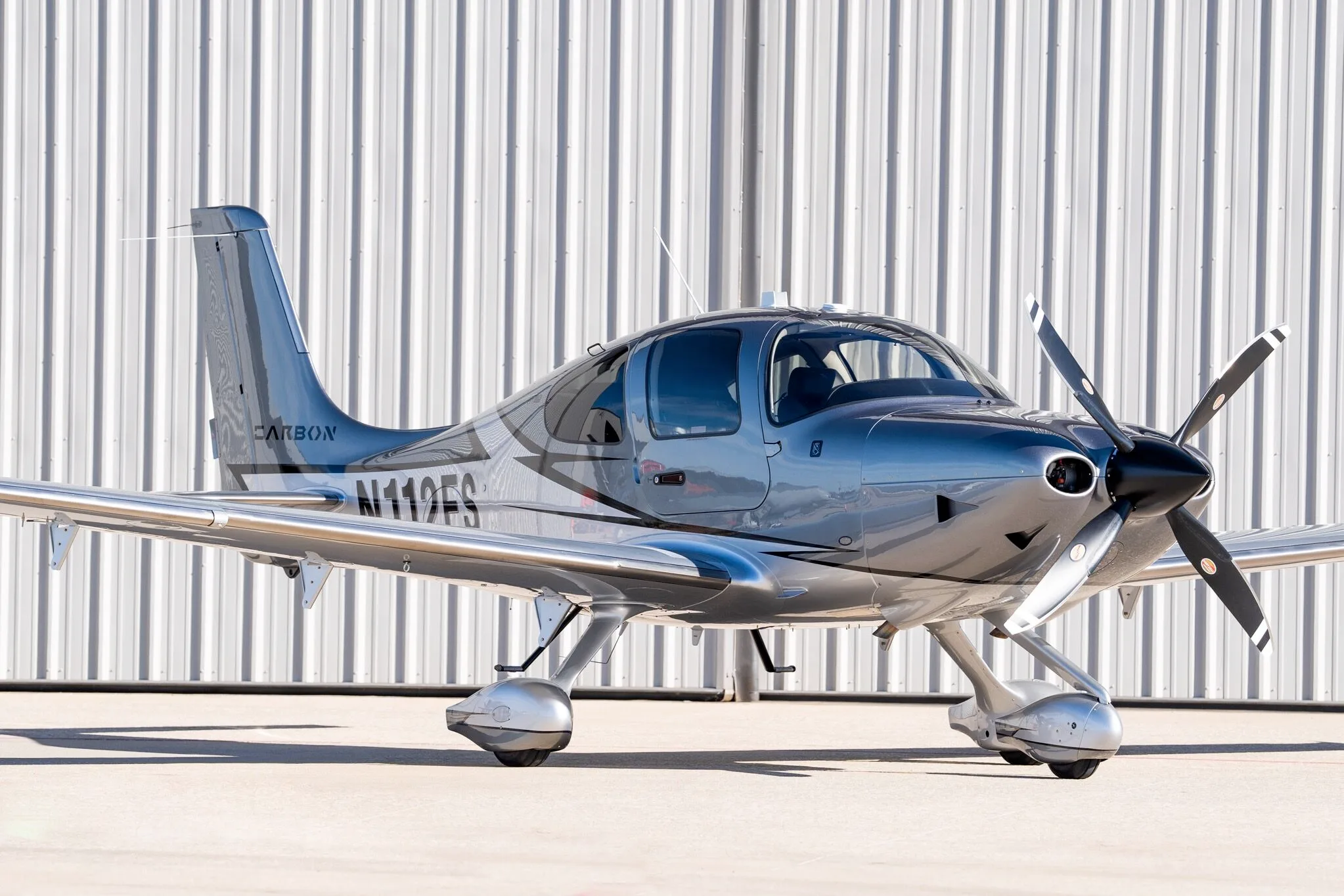 A front-right view of a silver and dark grey Cirrus SR22T Carbon Edition aircraft (N112FS) parked in front of a hangar.