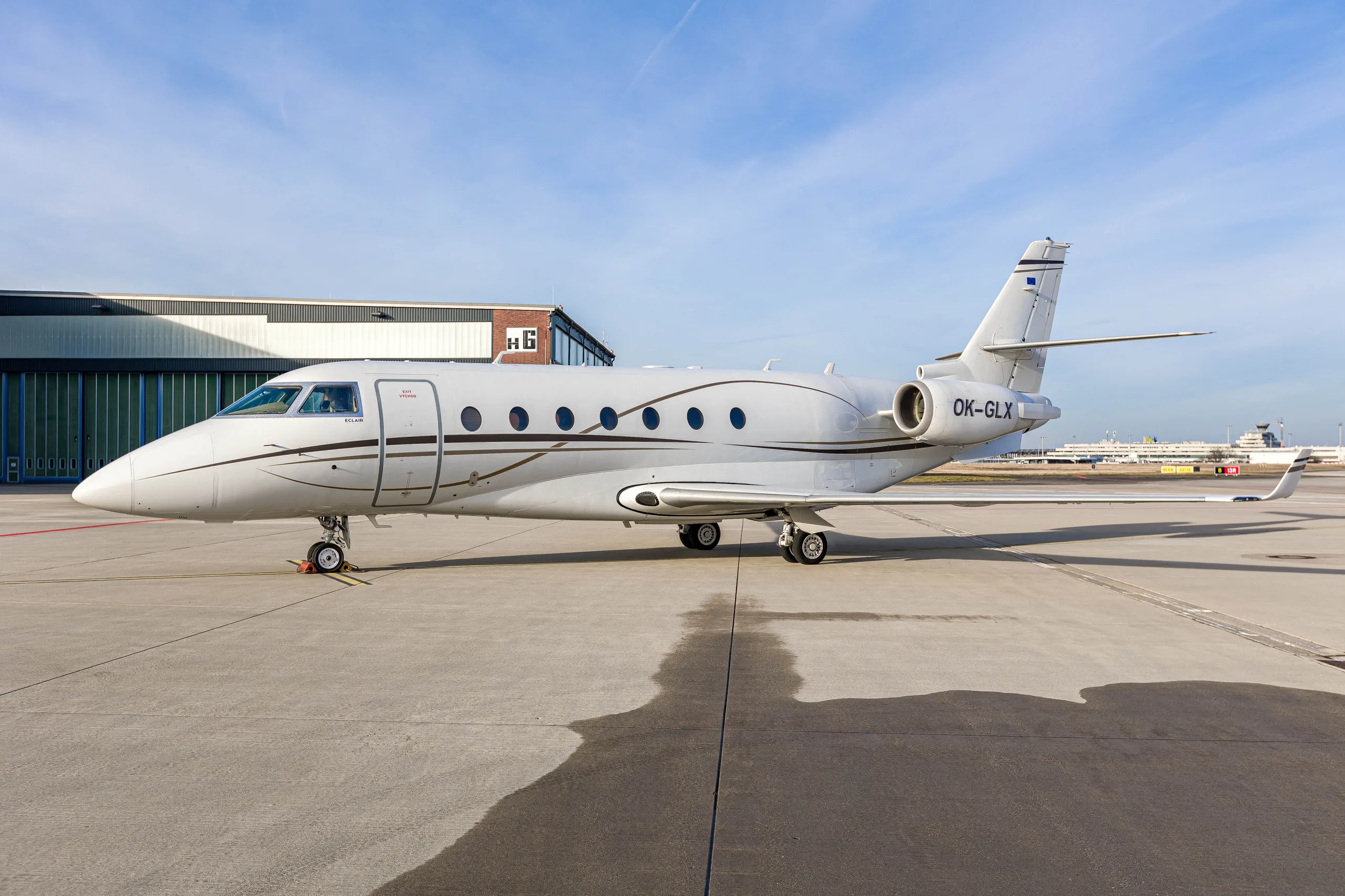 A white Gulfstream G200 business jet with brown accent stripes and registration OK-GLX parked on an airport tarmac.