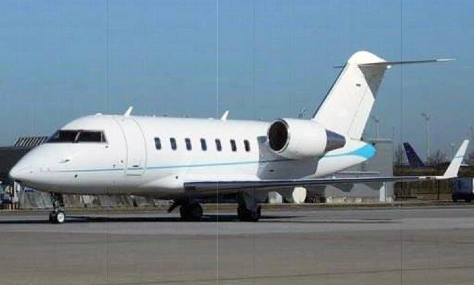 A white Bombardier Challenger business jet with a light blue stripe on its tail is parked on an airfield under a clear sky.