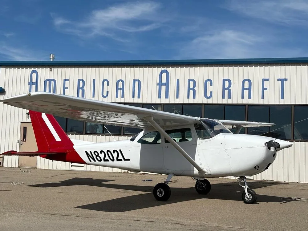A white Cessna 172 with a red tail, registration N8202L, parked on an tarmac in front of a building.