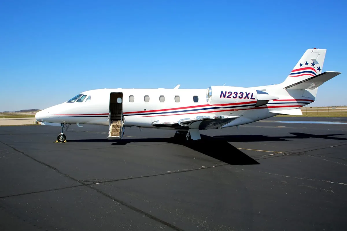 A white business jet with N233XL registration and patriotic tail stripes is parked on an airport tarmac with its door open.