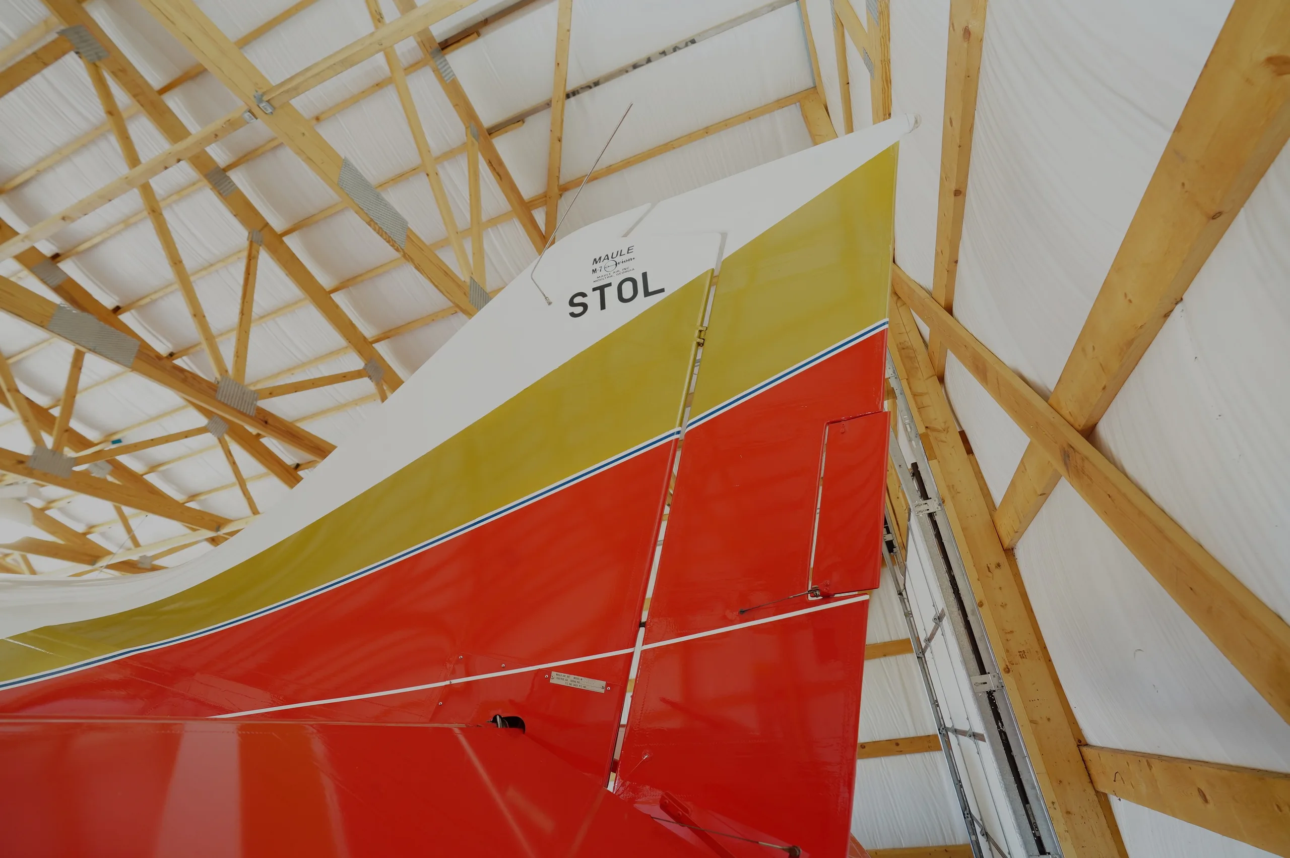 A close-up of the red, gold, and white tail of a Maule STOL aircraft inside a hangar.