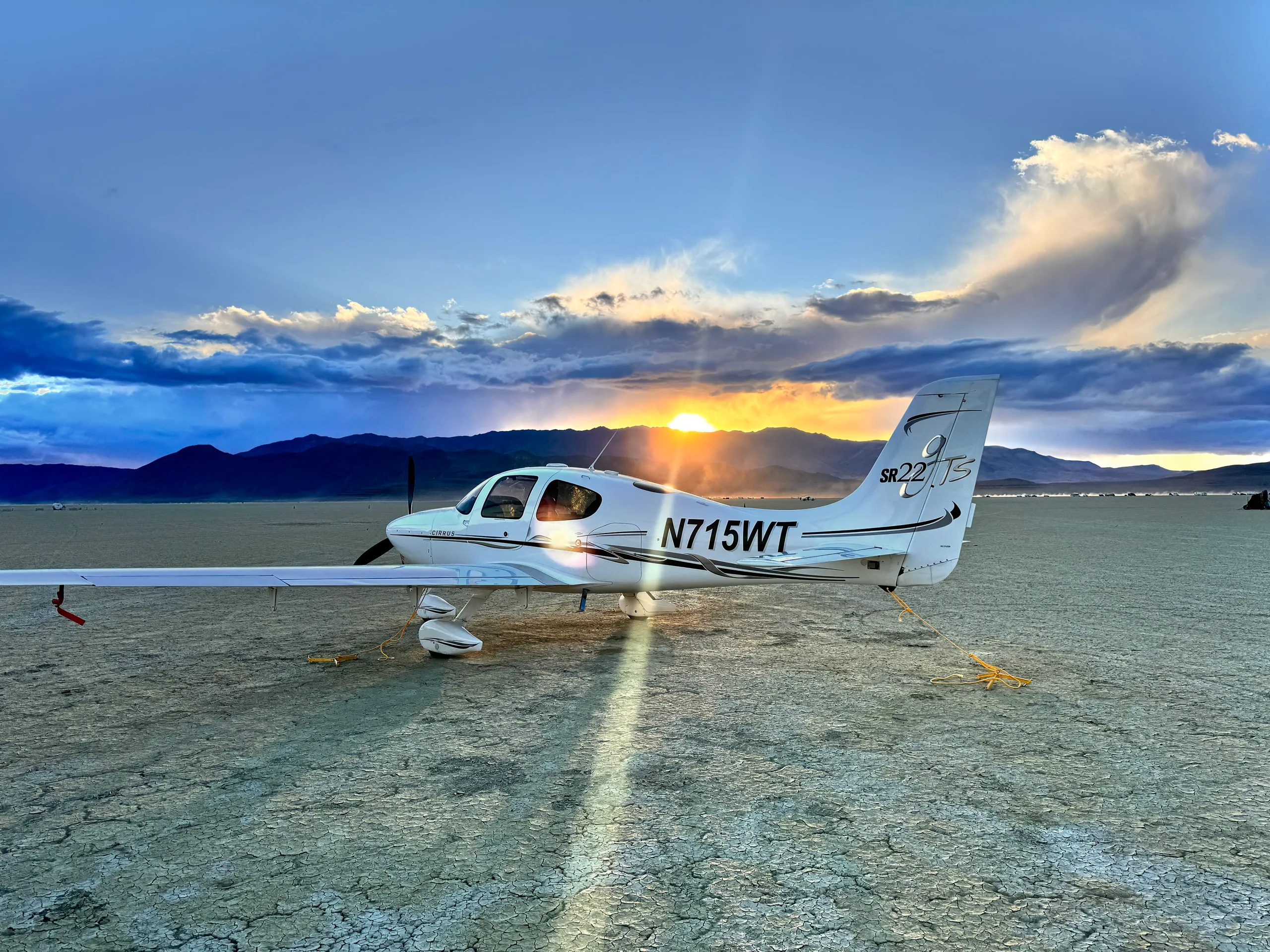 A white Cirrus SR22T aircraft (N715WT) parked on a cracked dry lake bed at sunset, with sun rays beaming from behind.