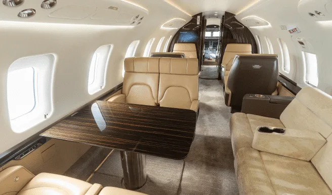 The luxurious interior of a business jet cabin with tan leather seats, a dark wood table, and ambient lighting.