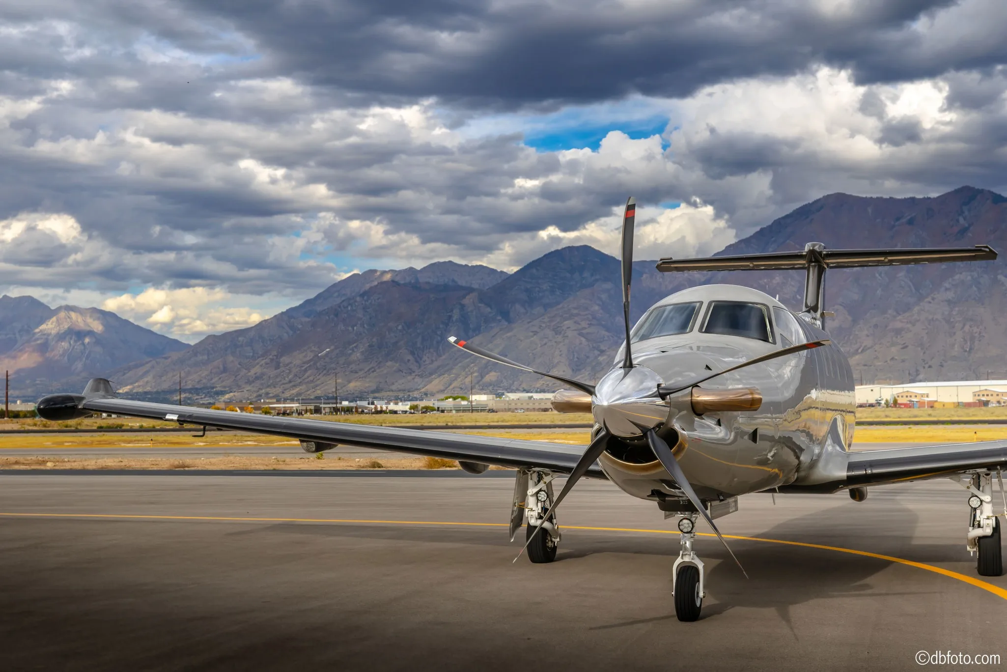 A silver Pilatus PC-12 turboprop aircraft is parked on an airport tarmac with mountains in the background under a dramatic cloudy sky.