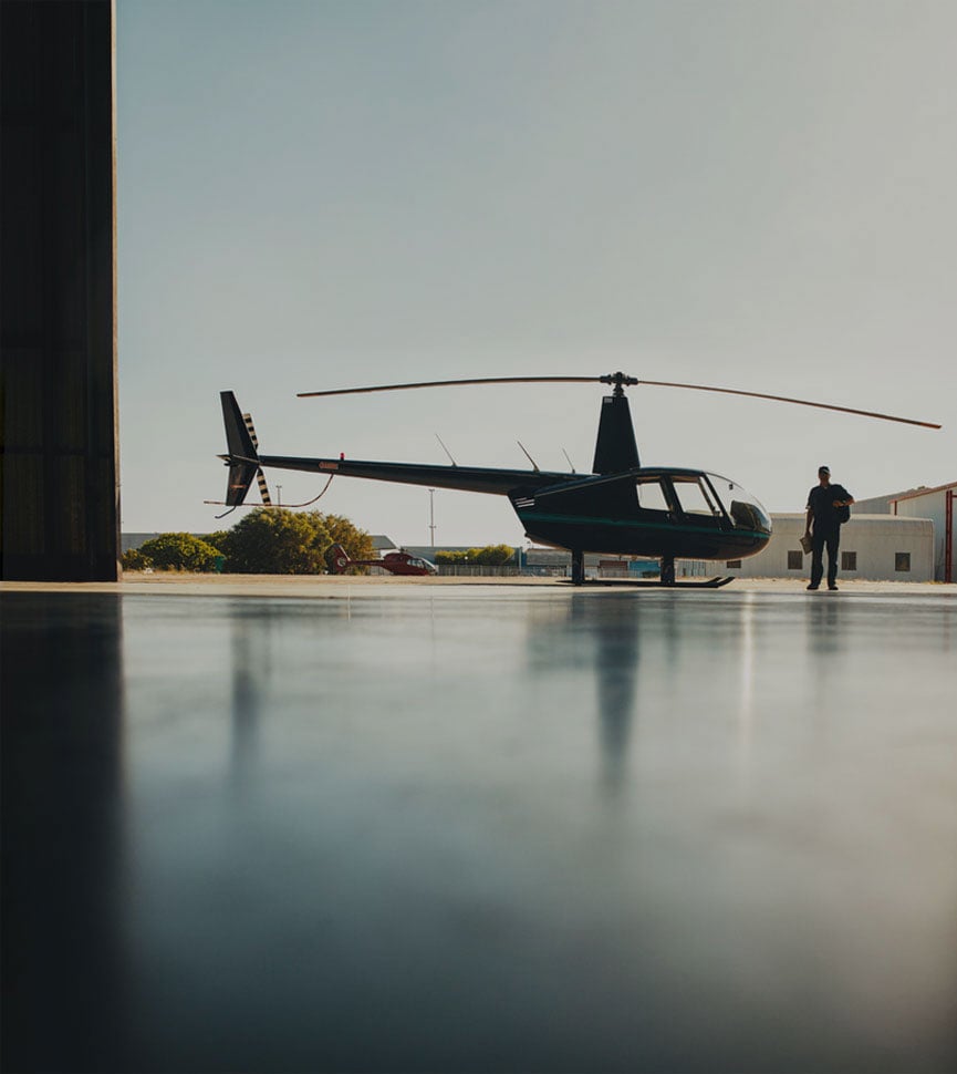 A black Robinson helicopter parked outside a hangar with a person standing nearby.